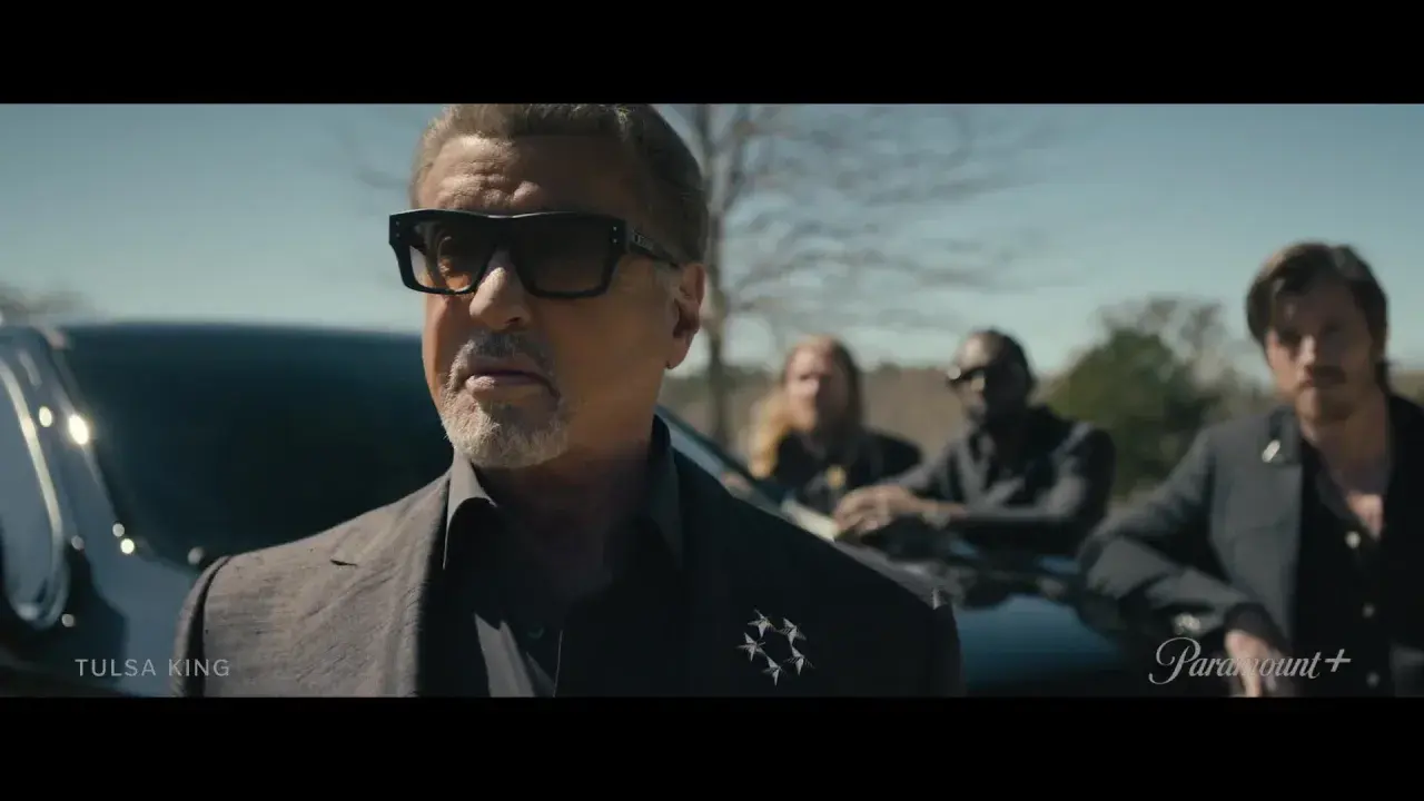 Sylvester Stallone als Tulsa King, mit Sonnenbrille und Anzug, steht vor einem Auto, begleitet von drei Männern.