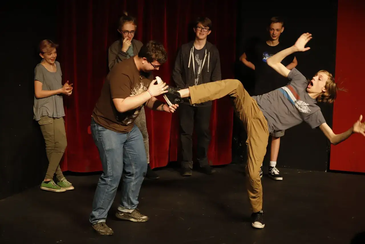 Junge Leute &uuml;ben beim Improvisationstheater Kurs eine Szene. Einer f&auml;llt dramatisch, w&auml;hrend die anderen zuschauen.