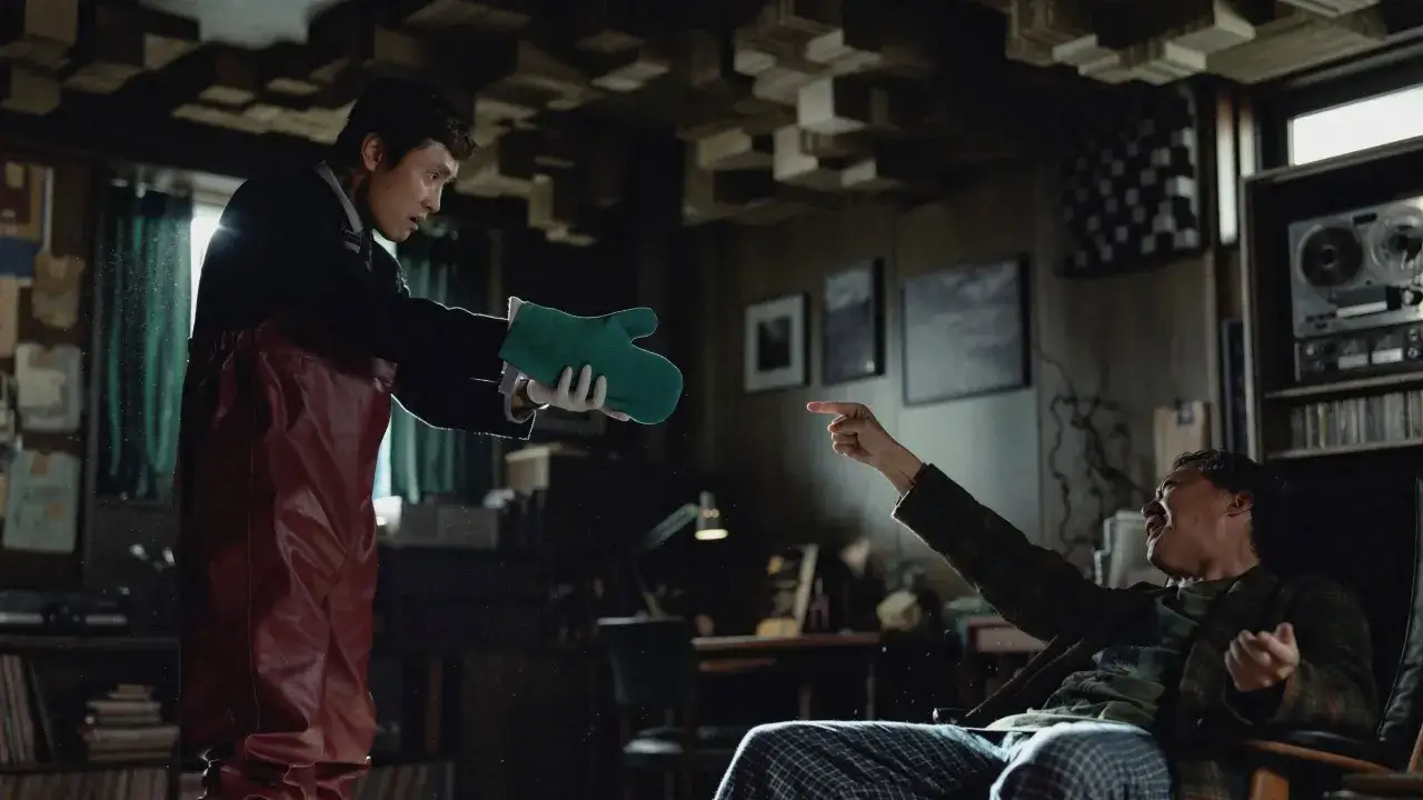 Zwei Männer in einem dunklen Raum, einer hält einen grünen Ofenhandschuh, der andere zeigt wütend darauf. Szene aus dem Film