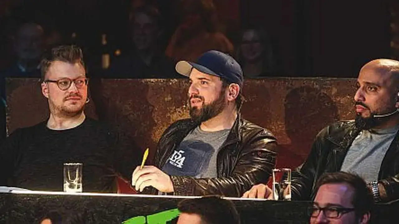 Drei M&auml;nner sitzen an einem Tisch. Der Mann links tr&auml;gt eine Brille. Der mittlere Mann mit Bart und Kappe h&auml;lt einen Stift. Das 