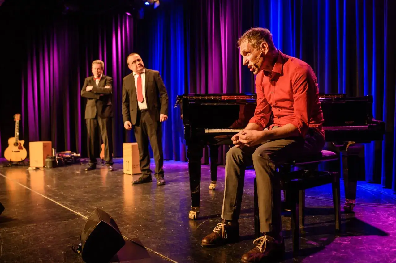 Bühnenperformance mit Klavier und drei Männern in Anzügen. Die Beleuchtung und die Szenerie vermitteln die Bedeutung von Kabarett: Unterhaltung mit Tiefgang.