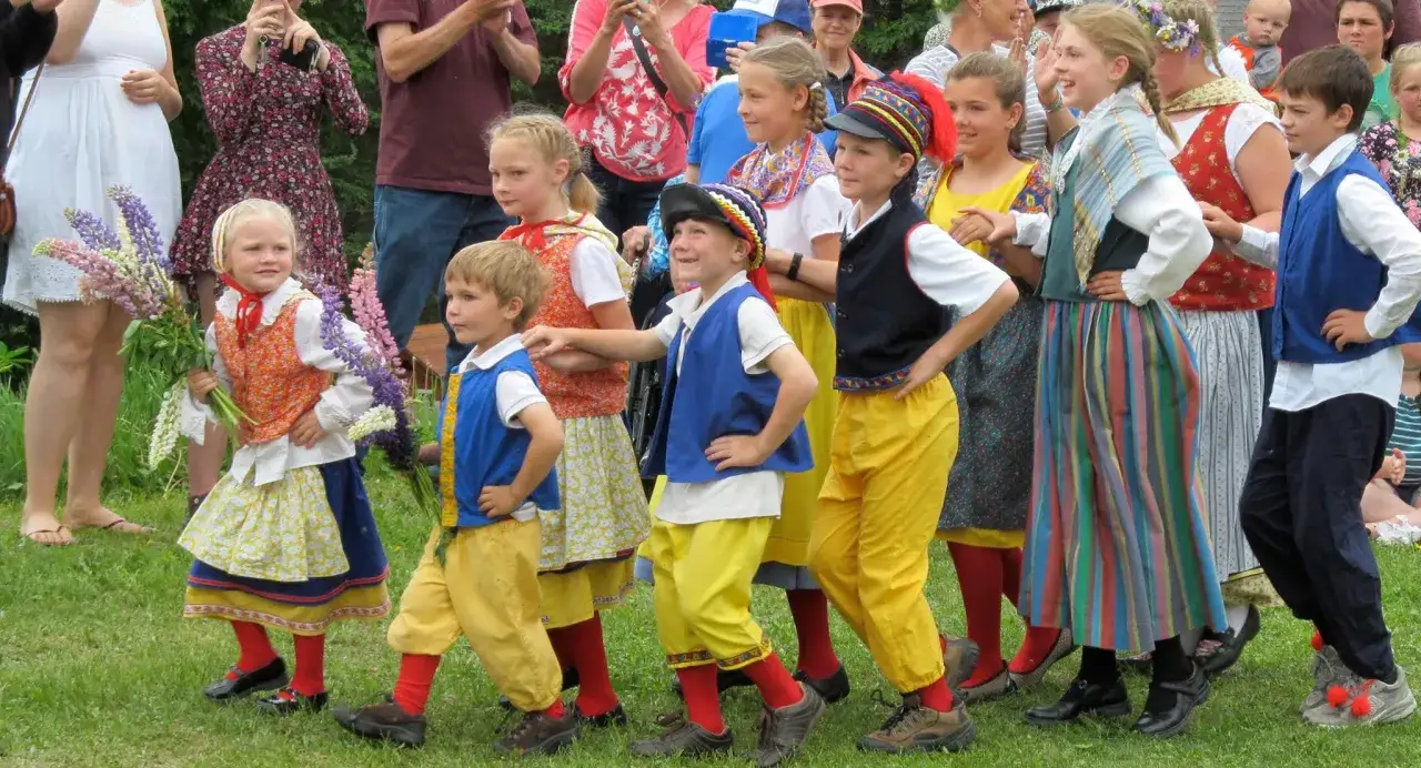 Kinder in traditioneller Kleidung, die an eine Midsommar-Besetzung erinnern, tanzen fröhlich. Bunte Blumen und Trachten prägen das Bild.