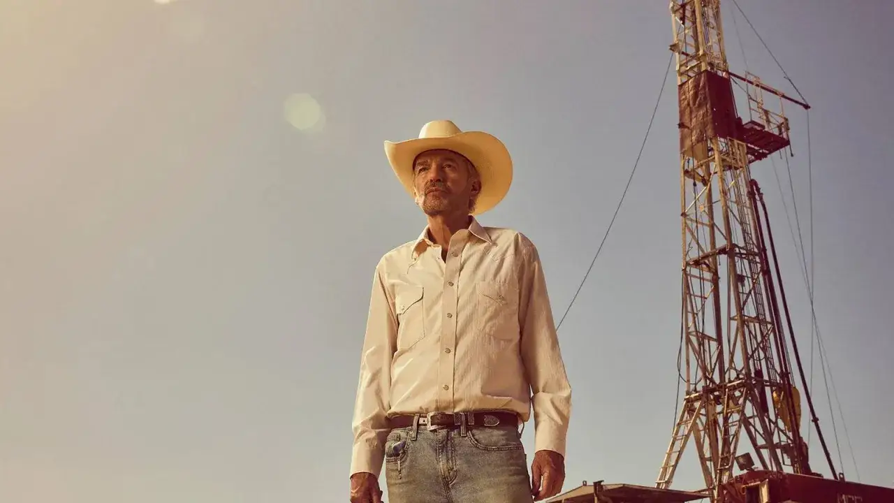 Ein Mann in Cowboyhut und Jeans steht vor einer Ölbohranlage. Die Szene erinnert an die