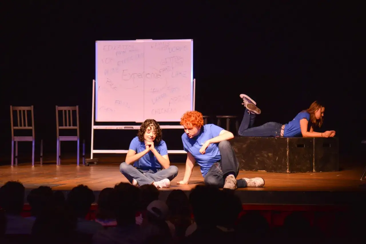 Jugendliche auf B&uuml;hne bei Improvisationstheater. Eine Frau liegt auf Kiste, zwei sitzen davor. Eine Tafel mit Begriffen wie 