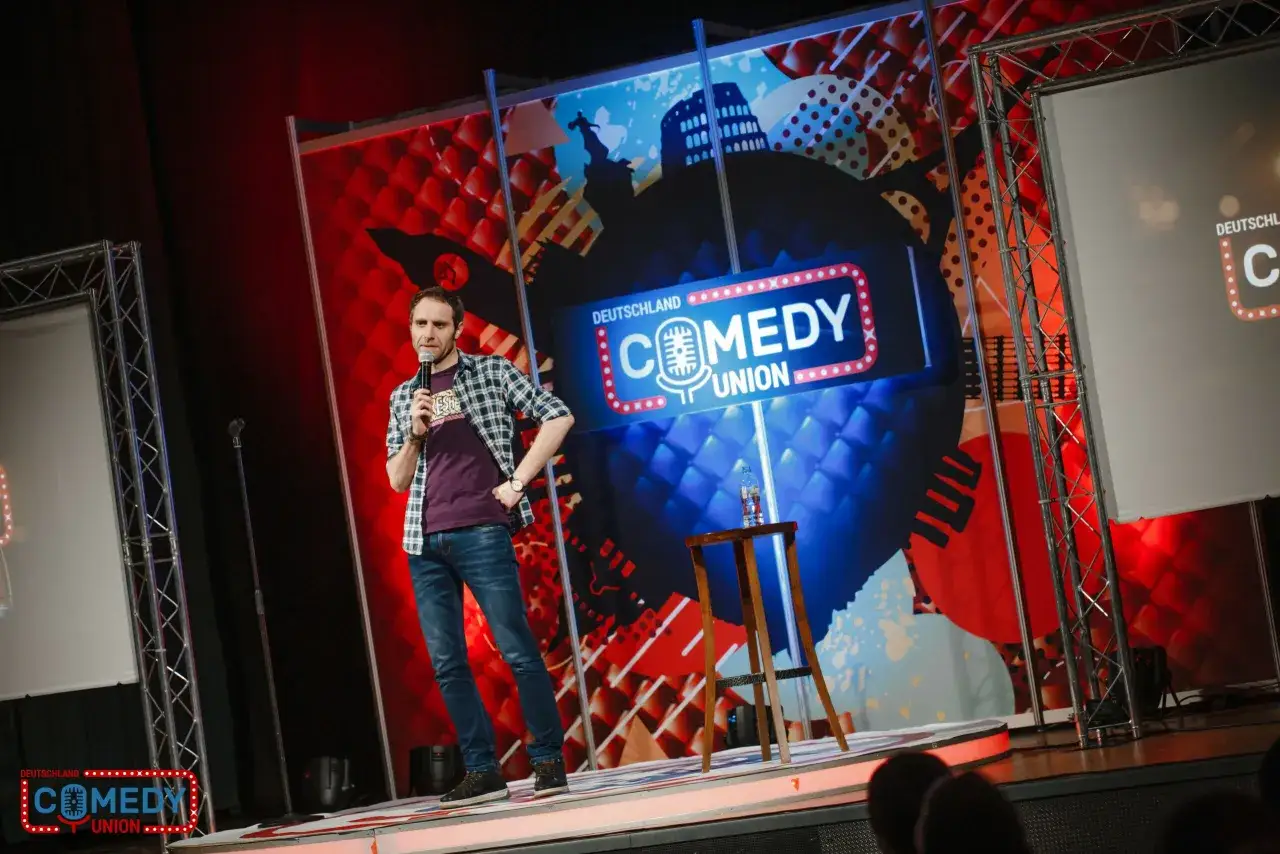 Ein Mann steht auf einer Bühne und hält ein Mikrofon. Er ist Teil einer stand up komödie.