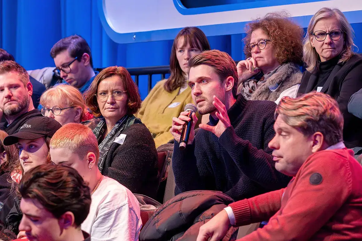 Ein Mann spricht ins Mikrofon, die Zuhörer sind gespannt. Kennste kennste, wer da wohl spricht?