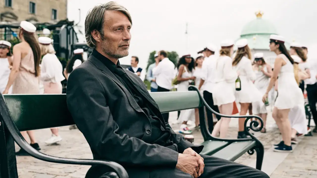 Mads Mikkelsen sitzt nachdenklich auf einer Bank, während im Hintergrund junge Leute in weißen Kleidern und Studentenmützen feiern. Ein Moment aus