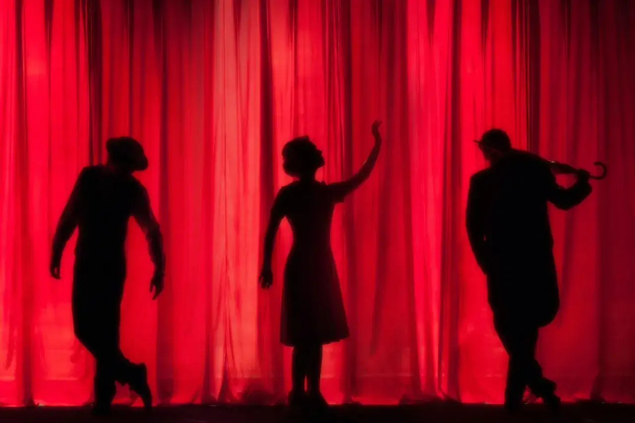 Drei Silhouetten vor rotem Vorhang. Ein Mann mit Hut, eine Frau mit erhobener Hand, ein Mann mit Regenschirm. Ein Szene wie im Brecht Theater.