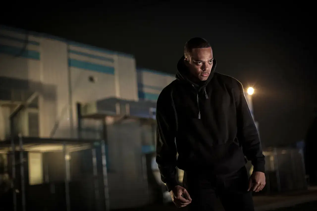 Ein Mann in einem schwarzen Hoodie steht im Dunkeln vor einem Geb&auml;ude. Die Szene erinnert an spannende Thriller-Filme.