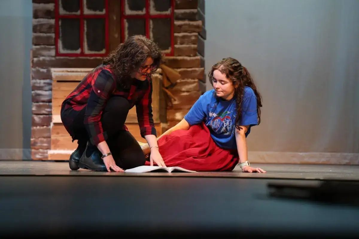 Zwei Frauen auf einer Bühne, eine kniet und zeigt auf ein Buch, die andere sitzt. Beispiele für Standbilder im Theater.