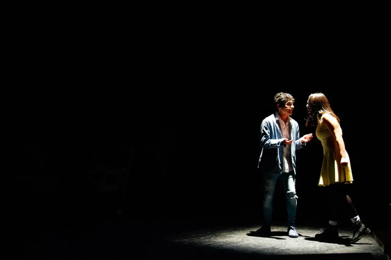 Zwei junge Schauspieler im Scheinwerferlicht. Ihre Gesten und Mimik deuten auf einen inneren Monolog, eine Definition von Gedanken, die nicht ausgesprochen werden.