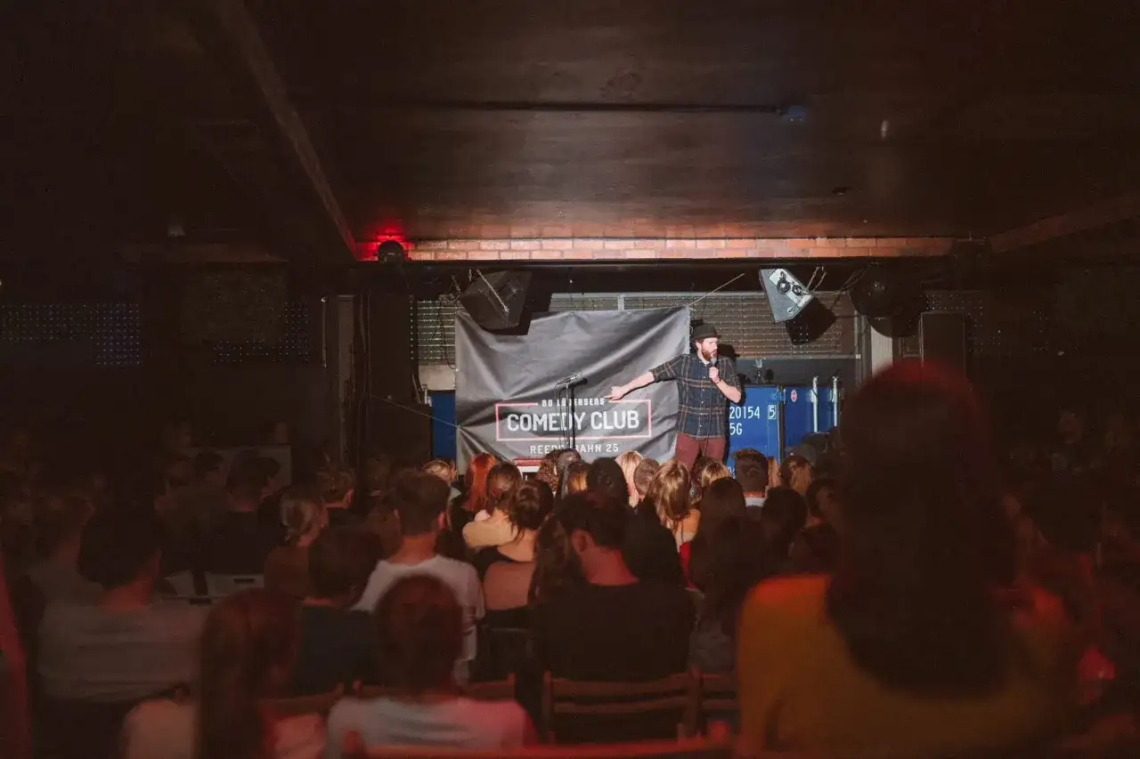 Ein Mann mit Hut und kariertem Hemd steht auf der Bühne eines Comedy Clubs und hält ein Mikrofon. Das Publikum sitzt im Dunkeln und lauscht gespannt dem open mic.