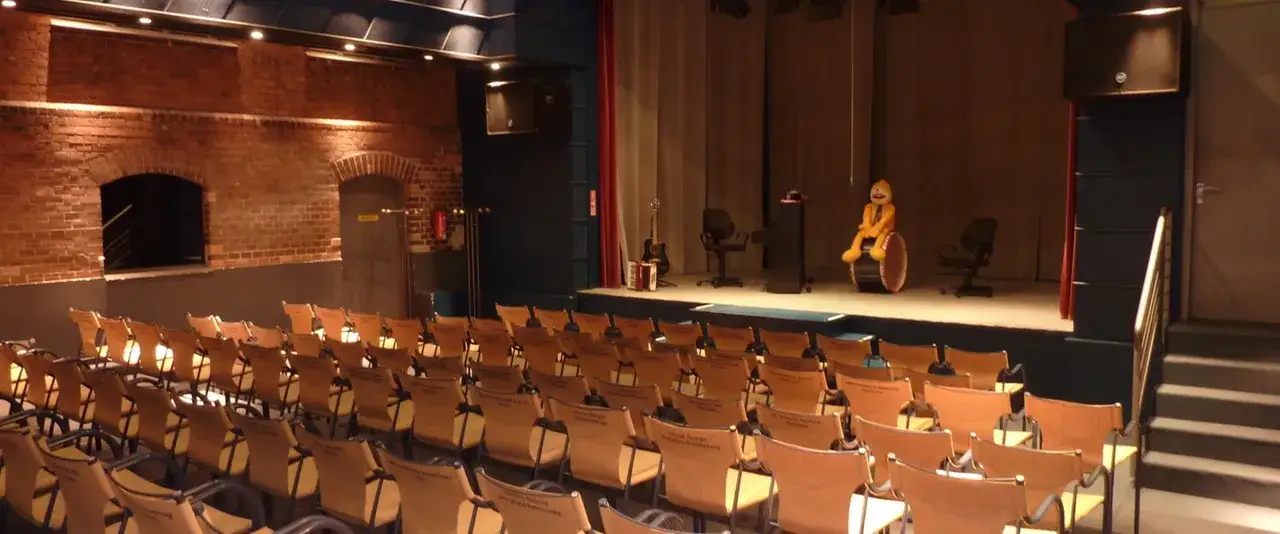 Bühne mit gelber Figur auf Trommel, Gitarre und Akkordeon. Die leeren Stuhlreihen lassen auf eine bevorstehende Kabarett-Vorstellung schließen.