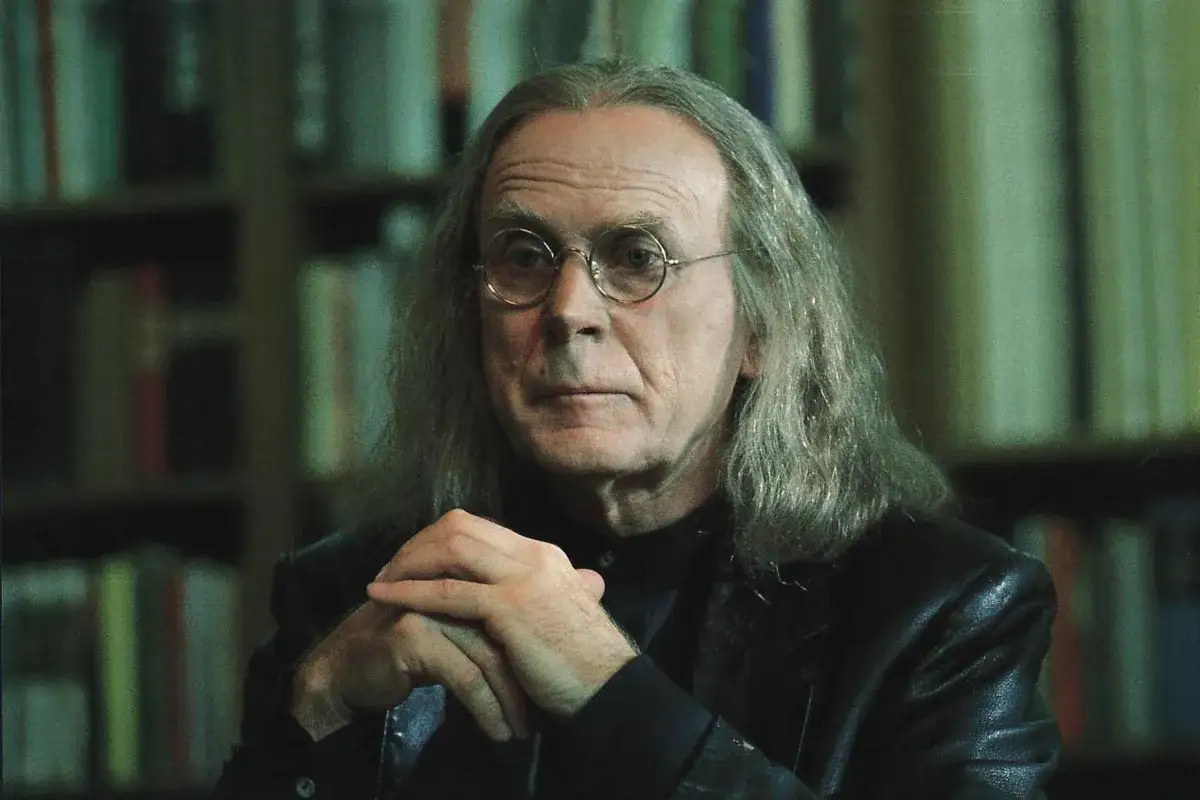 Ein Mann mit langen grauen Haaren und runder Brille, der an eine Loriot-Dichterlesung erinnert, sitzt vor einem Bücherregal.