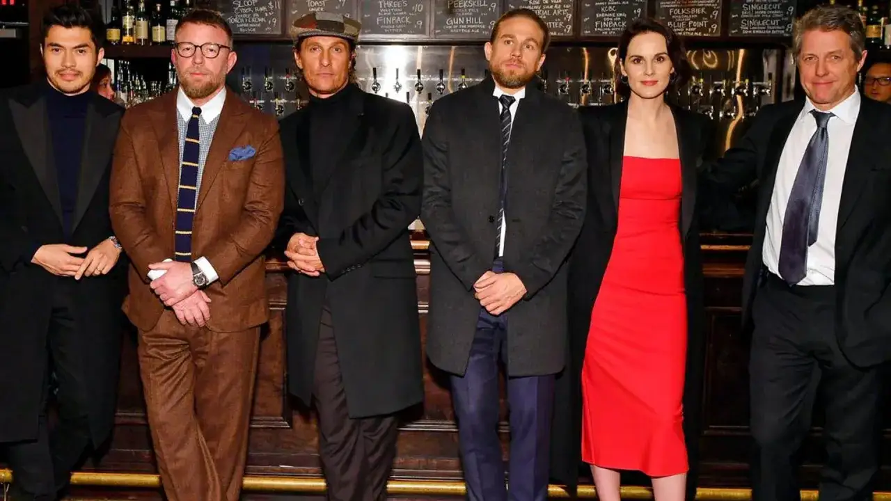Die Besetzung von The Gentlemen: Henry Golding, Colin Farrell, Matthew McConaughey, Charlie Hunnam, Michelle Dockery und Hugh Grant posieren vor einer Bar.