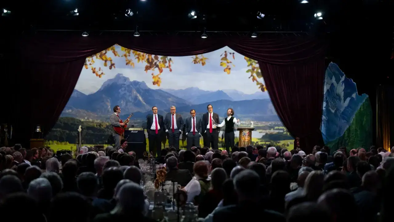 Fünf Männer in Anzügen und ein Gitarrist auf der Bühne. Die Kulisse zeigt Berge. Die Nockherberg-Kritik ist in vollem Gange.