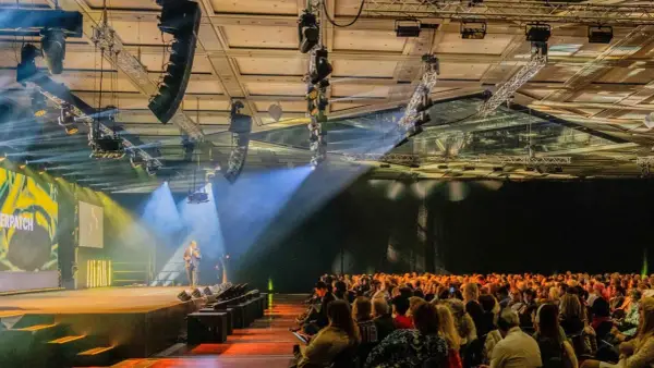 Bühne mit Redner, Publikum und Lichtshow. Die Atmosphäre erinnert an eine spontane Improvisation, Musik und ein inspirierendes Event.